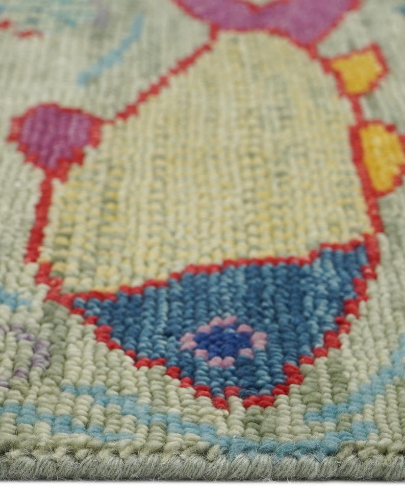 Op de afbeelding: Close-up van een handgetuft tapijt met een lichtgroene achtergrond. Het tapijt heeft een centraal ontwerp in geel, omlijnd in rood, met blauwe en roze accenten. De textuur van het tapijt is zichtbaar.