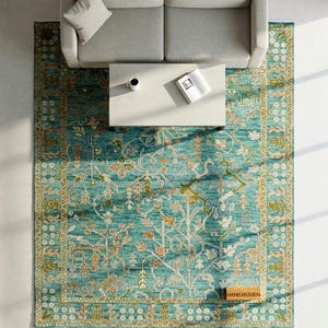 May include: Overhead view of a teal and gold area rug with an intricate floral pattern. A light gray sofa and white coffee table with a book and mug are above the rug. The rug has a rectangular shape and is placed on a white floor.