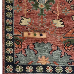 Puede incluir: Primer plano de una secci&oacute;n de una alfombra estampada con una muestra de color de madera. La alfombra presenta un fondo rojo con dise&ntilde;os florales verdes, azules y naranjas. Un borde azul oscuro enmarca el borde.