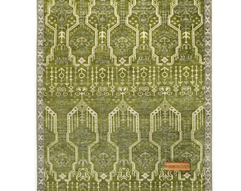 May include: A rectangular, olive green rug with a geometric pattern in off-white and gray. The rug features a central design of stylized shapes and a border with a repeating cross pattern. The tag reads "HANKWOVEN".