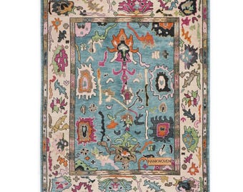 May include: Rectangular area rug with a teal center and a cream-colored border. The rug features an intricate pattern of colorful, abstract shapes in shades of pink, orange, green, and black. The rug is labeled "HANKWOVEN" in the lower right corner.
