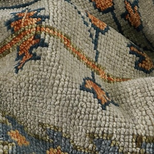 Puede incluir: Primer plano de una alfombra texturizada con un estampado floral. La alfombra presenta un fondo gris claro con acentos de naranja, azul y verde. El diseño incluye motivos florales estilizados y un borde.