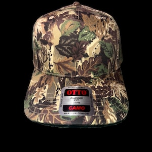 May include: A camouflage baseball cap with a curved brim. The cap features a leaf pattern in shades of green, brown, and tan. The front of the cap has a tag that reads "OTTO CAMO".