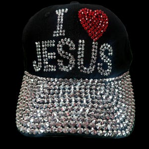 May include: Black baseball cap with the phrase "I LOVE JESUS" in rhinestones. A red rhinestone heart sits above "JESUS". The cap's brim is covered in silver rhinestones. A stylish accessory.