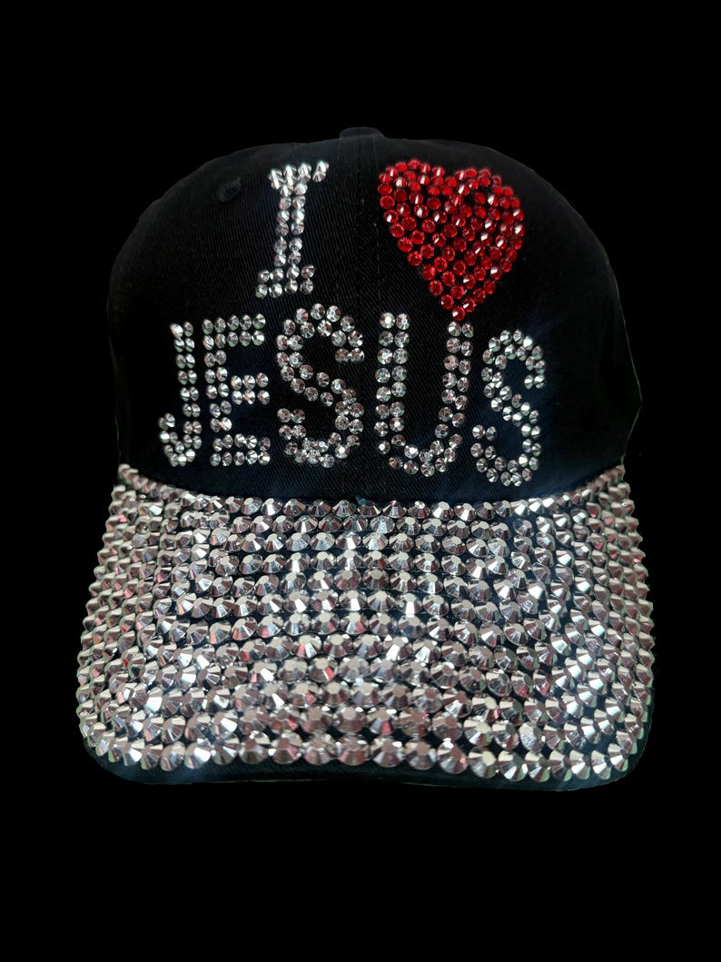 May include: Black baseball cap with the phrase "I LOVE JESUS" in rhinestones. A red rhinestone heart sits above "JESUS". The cap's brim is covered in silver rhinestones. A stylish accessory.