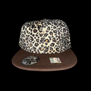 May include: A brown and tan leopard print baseball cap with a flat peak. The cap features a brown peak and a leopard print crown. The cap has a Sports Authentic logo on the peak.