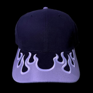 May include: A navy blue baseball cap with a lavender flame design on the brim. The cap has a curved brim and a structured crown. The flame design is embroidered in a lighter shade of lavender.