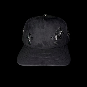 Puede incluir: Gorra de béisbol negra con adornos de cruces. La gorra presenta múltiples cruces decorativas en la parte delantera y los laterales. La visera tiene un diseño curvo con detalles cosidos. La gorra está hecha de un material suave similar al ante.