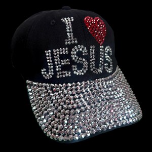 May include: Black baseball cap with the text "I ❤️ JESUS" in silver rhinestones and a red rhinestone heart. The brim is fully covered in silver rhinestones. A stylish and faith-based accessory.