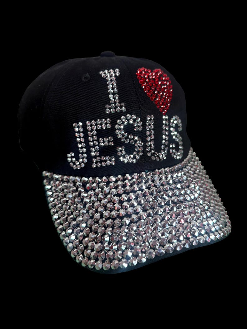 May include: Black baseball cap with the text "I ❤️ JESUS" in silver rhinestones and a red rhinestone heart. The brim is fully covered in silver rhinestones. A stylish and faith-based accessory.
