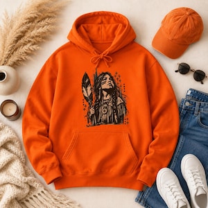 Op de afbeelding: Een feloranje hoodie met een zwarte grafische print van een persoon met lang gevlochten haar die een veer vasthoudt. De trui is voorzien van een kangoeroezak aan de voorzijde en bijpassende trekkoorden voor de capuchon.