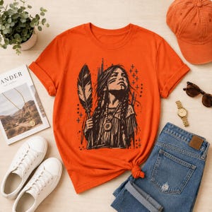 Op de afbeelding: Een oranje t-shirt met korte mouwen en een zwarte grafische illustratie van een persoon met lang haar, gekleed in een top met franjes en een ketting, die een grote veer vasthoudt. Het ontwerp bevat kleine kruisjes en verticale lijnen.