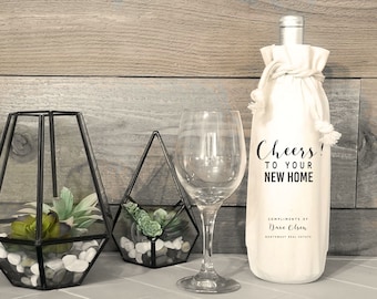 Może przedstawiać: Biały worek na butelkę wina z czarnym napisem: "Cheers! To your new home. Compliments of Dave Olsen, Northwest Real Estate". Worek jest związany białym sznurkiem i leży na drewnianej powierzchni z dwoma szklanymi terrariami i kieliszkiem do wina.