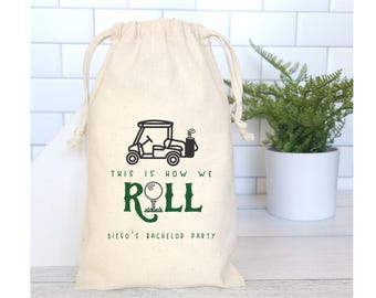May include: A beige drawstring bag with a golf cart and golf ball design. The text "THIS IS HOW WE ROLL" and "DIEGO'S BACHELOR PARTY" are printed in black and green. The bag is made of a natural fabric.