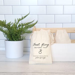 May include: A white cotton drawstring bag with the text "Last Fling Before The" and a diamond ring icon. The text "Meghan's Bachelorette Palm Springs" is also printed on the bag.