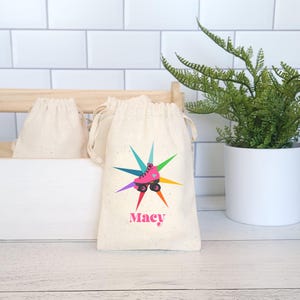 May include: A white cotton drawstring bag with a colorful starburst design and a pink roller skate with a heart on it. The bag has the name "Macy" printed on it.