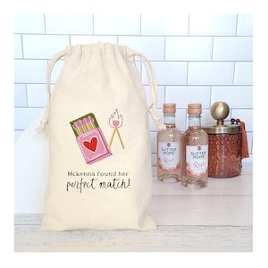 May include: A beige drawstring bag with a pink matchbox and a burning match graphic. The text "McKenna found her perfect match!" is printed on the bag. Two bottles of Sutter Home Rosé wine and a decorative jar are next to the bag.