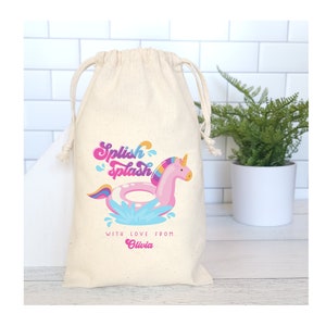 May include: A white cotton drawstring bag with a pink unicorn floating on a blue and pink inflatable ring. The bag has the text "Splish Splash" and "With love from Olivia" printed on it.
