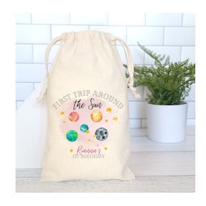 May include: A white cotton drawstring bag with a colorful graphic of planets and stars. The text on the bag reads "First Trip Around the Sun" and "Rianna's 1st Birthday".