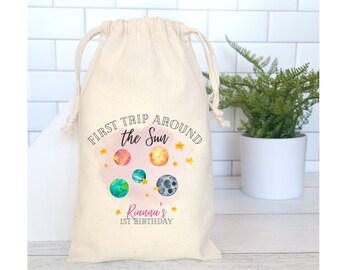 May include: A white cotton drawstring bag with a colorful graphic of planets and stars. The text on the bag reads "First Trip Around the Sun" and "Rianna's 1st Birthday".