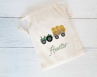 May include: A white cotton drawstring bag with a green tractor pulling a wagon full of hay bales. The bag has the name "Hunter" written on it in green.