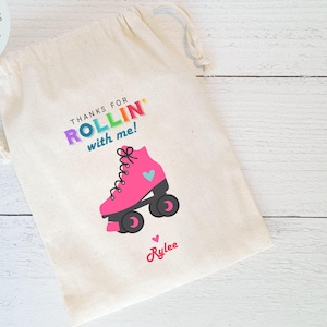May include: A white cotton drawstring bag with the text "Thanks for Rollin' with me!" in rainbow letters. A pink roller skate with blue wheels and a blue heart is pictured below the text. The name "Rylee" is written in pink cursive below the roller skate.