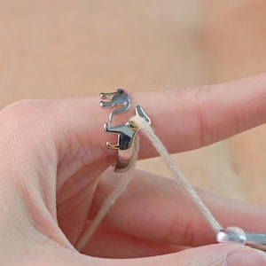 May include: A silver-colored ring with a dachshund dog design. The ring features a detailed depiction of the dog's head and body. The ring is shown on a finger, with a light-colored string looped through it.