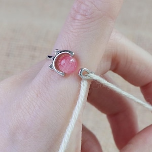 Könnte beinhalten: Ein silberner Ring mit Katzenohren-Design, der einen rosa Edelstein hält. Ein weißes Garn ist an einem silbernen Haken befestigt. Der Ring befindet sich an einem Finger, und das Garn wird gehalten.