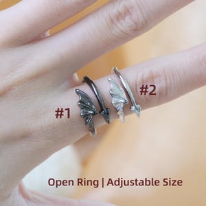 May include: Four adjustable open rings are displayed, two with a dark finish and two with a silver finish. Each ring features a wing design. The text "Open Ring | Adjustable Size" is visible.