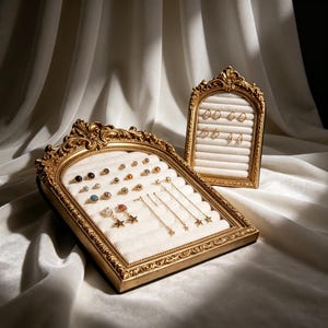 以下が含まれることがあります： クリーム色のベルベットインサートが付いた、装飾的な金色のアーチ型ジュエリーディスプレイフレーム2点。大きい方のフレームには様々なスタッドピアスや星型のぶら下がりピアスが並び、小さい方の直立したフレームには小さな金のフープピアスが飾られています。