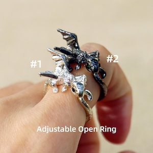 May include: Two dragon-shaped rings, one silver and one black, are displayed. The silver ring is detailed with small embellishments. The black ring has a sleek, polished appearance. The rings are adjustable and open, as indicated by the text in the image.
