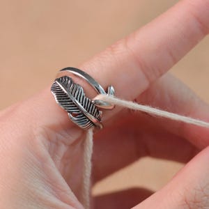 Pode incluir: Um anel de pena prateado com detalhes intrincados. O anel apresenta um design de pena detalhado na frente e uma banda lisa que envolve o dedo. Um fio branco é passado pelo anel.