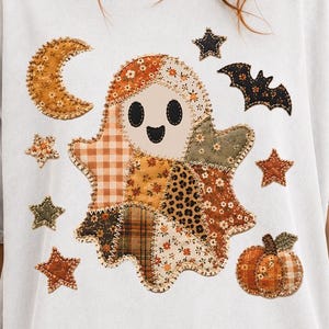 May include: A light gray short-sleeve t-shirt featuring a central graphic of a ghost made of various patchwork fabric patterns. The design includes a crescent moon, stars, a bat, and a pumpkin, all rendered in earthy autumn tones of orange, brown, and green.