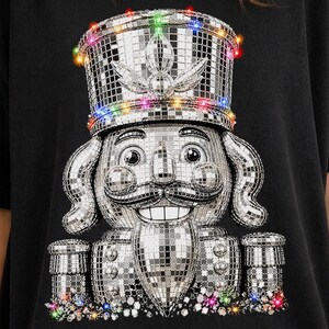 May include: A black t-shirt featuring a graphic of a nutcracker made of silver disco ball mirror tiles. The nutcracker wears a tall hat adorned with small, colorful glowing lights, and the base is decorated with a cluster of vibrant, multi-colored gems.