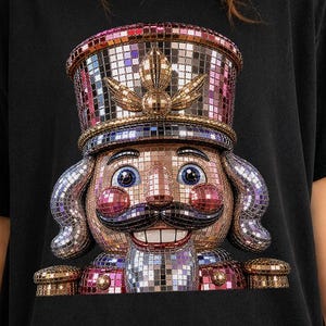 May include: A black t-shirt featuring a large graphic of a nutcracker made of reflective, multi-colored disco ball mirror tiles. The nutcracker has a tall hat, a prominent mustache, and a wide smile, rendered in shades of pink, silver, and purple.