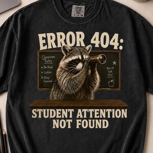 May include: A black short-sleeve t-shirt featuring a graphic of a raccoon looking through a telescope in front of a chalkboard. The text reads ERROR 404: STUDENT ATTENTION NOT FOUND, with classroom rules listed on the board next to the raccoon.