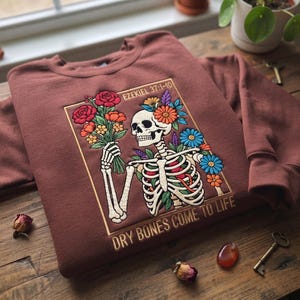 May include: A maroon crewneck sweatshirt featuring an embroidered graphic of a skeleton holding a bouquet of colorful flowers. The design includes the text Ezekiel 37:1-10 and Dry Bones Come To Life, framed within a rectangular gold border.