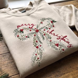 May include: A beige crewneck sweatshirt featuring an embroidered design of a bow made from pine branches and red berries. The text "merry christmas" is stitched in red cursive script across the center of the bow design.