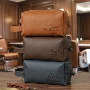 May include: Three leather toiletry bags stacked on a wooden surface. The top bag is brown with "William BEST MAN" engraved. The middle bag is dark brown with a deer antler design and "JAMES GROOMSMAN" engraved. The bottom bag is black with "Justin GROOMSMAN" engraved.