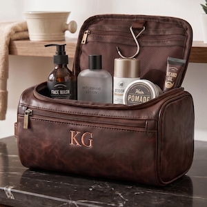 May include: A dark brown leather wash bag with the initials KG embossed on the front. The bag is open, revealing a metal hanging hook and several grooming products, including face wash, aftershave lotion, deodorant, hair pomade, and beard oil.