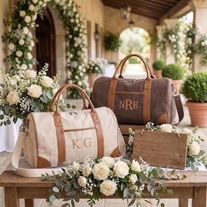 May include: Two canvas duffel bags with tan leather trim and handles. One bag is cream-colored with the monogram KG, and the other is dark brown with the monogram NRE. Both bags feature a zippered top closure and a detachable shoulder strap.