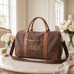 May include: A brown canvas duffel bag with tan leather trim, featuring the embroidered monogram NRE in gold thread on the front pocket, two top handles, and a detachable dark brown shoulder strap with metal hardware and zipper closures.
