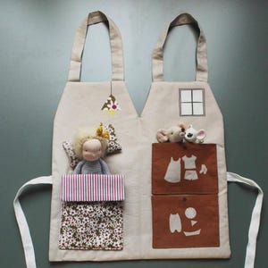Puede incluir: Delantal de tela beige con dos bolsillos y un diseño en forma de casa. El delantal presenta una muñeca con una almohada, una manta a rayas y dos ratones de juguete. Los bolsillos tienen ilustraciones de ropa y notas musicales.
