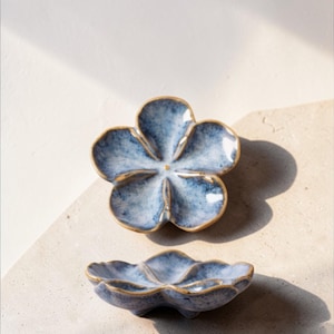 May include: Two blue ceramic flower-shaped dishes with a gold rim. The dishes have a mottled blue glaze. One dish is positioned above the other, both resting on a light-coloured surface.