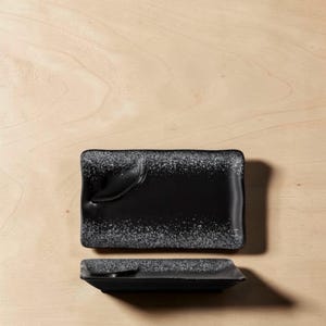 May include: Two rectangular black ceramic serving dishes. The dishes have a glossy finish with a speckled, white border. One dish is positioned above the other, with a slight overlap, on a light wood surface.