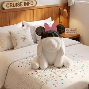 May include: A white decorative dog figurine with black ears and a pink bow, sitting on a bed with white linens. The bed has pillows with anchor designs. The headboard has a sign that says "CRUISE INFO".