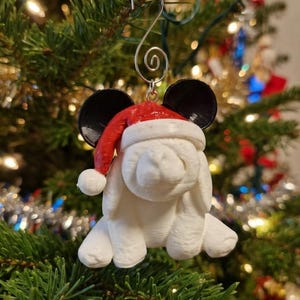 May include: A white 3D printed rabbit ornament featuring black mouse-like ears and a red and white Santa hat. The figure is suspended by a silver metal spiral hook. The rabbit has long drooping ears and a textured surface finish throughout the piece.