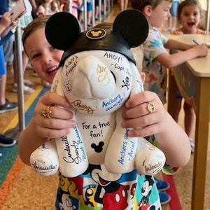 May include: A white Mickey Mouse-shaped plush toy with black ears and a black hat, covered in signatures and phrases like "AHOY!" and "For a true fan!". The toy is held by a person wearing a colorful Mickey Mouse-themed shirt.