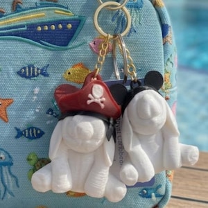 May include: Two white bunny keychains with black ears, one wearing a red pirate hat with a skull and crossbones. The keychains hang from a gold ring. The background is a light blue bag with an embroidered cruise ship.