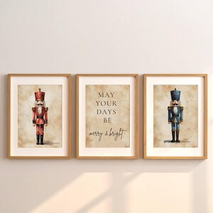May include: Three framed prints with a Christmas theme. Two prints feature watercolor nutcracker soldiers in red and blue uniforms. The centre print has the text "MAY YOUR DAYS BE merry & bright" in black script on a beige background.